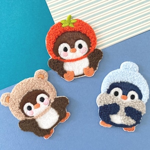 Op de afbeelding: Drie geborduurde pinguïnpatches. Eén pinguïn draagt een tomatenhoed, een andere een berenhoed en de derde een blauwe wintermuts. Elke pinguïn heeft een witte buik, bruine vleugels en oranje voeten. De achtergrond is blauw en wit.
