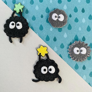 May include: Embroidered patches of cartoon characters. Two black characters with white eyes and star-shaped details, and two grey, round characters with white eyes. The patches are on a blue and white background.