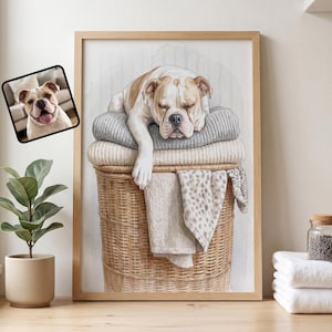 May include: A framed wall art print featuring a watercolor illustration of a white and tan bulldog sleeping on a stack of folded gray and beige blankets inside a woven wicker laundry basket, with a small inset photo of the same dog.