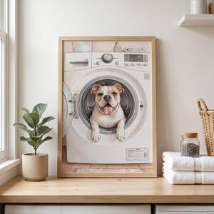 May include: A framed wall art print featuring a light-colored bulldog with its head and front paws poking out of the open circular door of a white front-loading washing machine. The dog has a happy expression with its tongue hanging out.