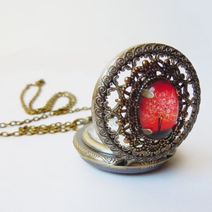 Puede incluir: Un reloj de bolsillo de bronce con un diseño de árbol rojo y blanco en la esfera. El reloj tiene un diseño de filigrana alrededor del borde y está unido a una cadena.