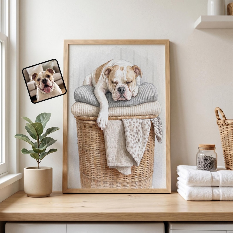 May include: A framed wall art print featuring a watercolor illustration of a white and tan bulldog sleeping on a stack of folded gray and beige blankets inside a woven wicker laundry basket, with a small inset photo of the same dog.