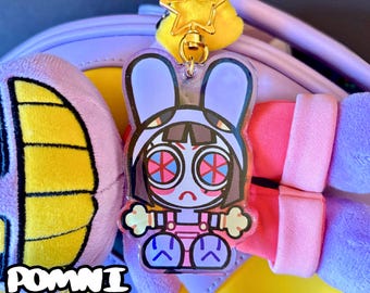 May include: A clear acrylic Pomni keychain featuring a cartoon rabbit character with large, spiral eyes and a sad expression. The keychain has a gold star charm and the text "+ STICKERS" is visible. The words "POMNI KEYCHAIN" are also displayed.