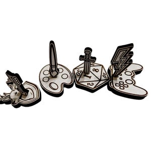 May include: A collection of wooden cutouts featuring various designs. Included are a deer head, a paintbrush on a palette, a sword on a die, and a lightning bolt on a game controller. Each cutout has a black outline.