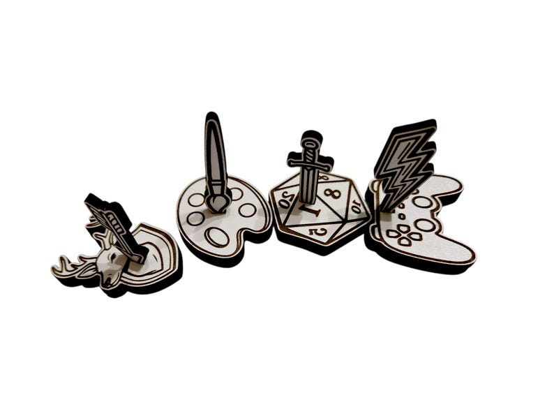 May include: A collection of wooden cutouts featuring various designs. Included are a deer head, a paintbrush on a palette, a sword on a die, and a lightning bolt on a game controller. Each cutout has a black outline.