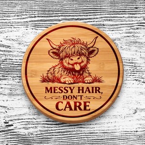 May include: A round bamboo cutting board featuring a laser-engraved illustration of a shaggy Highland cow with its tongue sticking out. The text "MESSY HAIR, DON'T CARE" is engraved below the cow, all enclosed within a circular border line.