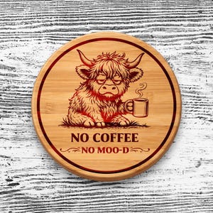 May include: A round bamboo coaster featuring a dark red engraved illustration of a shaggy Highland cow holding a coffee mug. The text NO COFFEE NO MOO-D is printed below the cow, all enclosed within a dark red circular border on the wood.