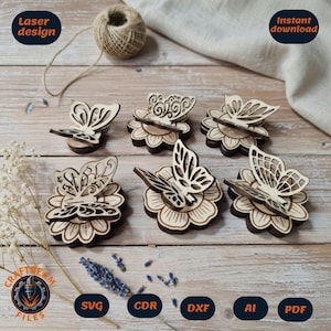 May include: Six laser-cut wooden butterfly ornaments, each featuring intricate wing patterns and mounted on a circular flower-shaped base. The natural light-colored wood pieces are arranged on a wooden surface with a ball of twine and dried flowers.