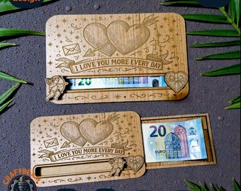 May include: Two rectangular wooden money holders featuring laser-engraved designs of two hearts pierced by an arrow, a small envelope, and a cherub. The text I LOVE YOU MORE EVERY DAY is engraved on the front. One holder contains a 20 Euro bill.