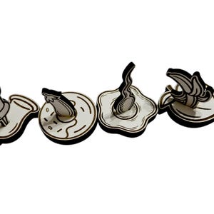 May include: Four wooden cutouts in a row, each with a black outline and white interior. The shapes include a pushpin, donut, fried egg, and banana. The designs are simple and cartoon-like, with a focus on food and office supplies.