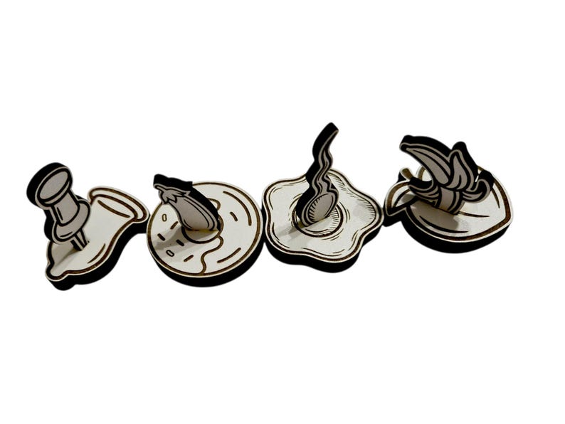 May include: Four wooden cutouts in a row, each with a black outline and white interior. The shapes include a pushpin, donut, fried egg, and banana. The designs are simple and cartoon-like, with a focus on food and office supplies.