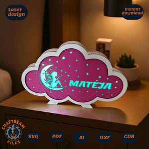 Könnte beinhalten: Ein wolkenförmiges Nachtlicht mit rosa Innenraum und weißem Rahmen. Das Design zeigt einen Mond mit einer Fee und dem Namen "MATEJA" in Türkis. Das Licht steht auf einem Nachttisch aus Holz.