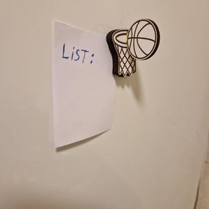 May include: A white piece of paper with the word "List:" written in blue ink is held in place by a basketball-themed magnet. The magnet features a basketball and hoop design with black and white details.
