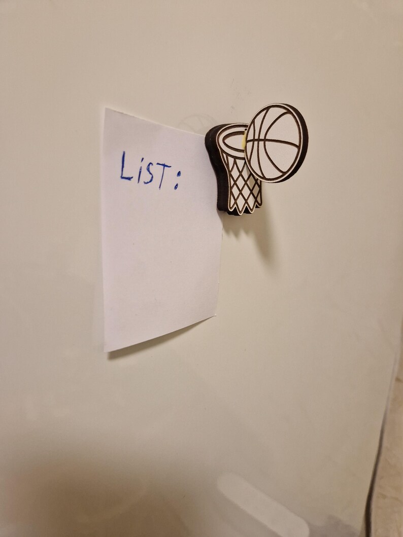 May include: A white piece of paper with the word "List:" written in blue ink is held in place by a basketball-themed magnet. The magnet features a basketball and hoop design with black and white details.