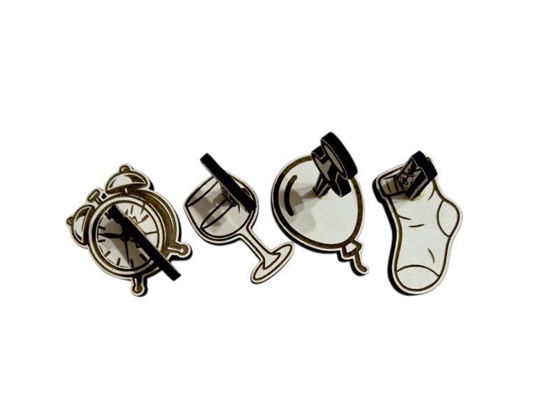 May include: A collection of four wooden cutouts: an alarm clock, a wine glass, a balloon, and a sock. Each cutout has a black accent, creating a contrast against the light wood. The clock hands are black, and the wine glass has a black stem.