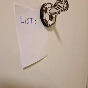May include: A wooden magnet featuring a soccer ball and a soccer cleat. The magnet holds a white piece of paper with the word "List:" written in blue ink. The magnet is attached to a white surface.