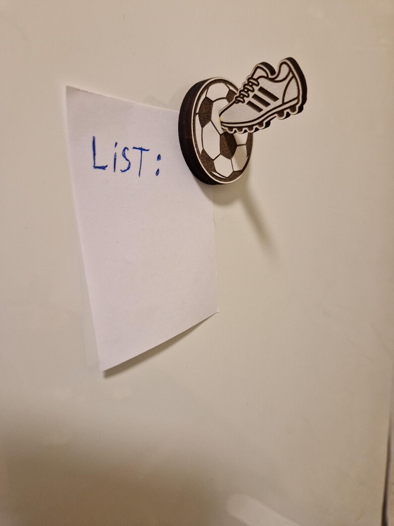 May include: A wooden magnet featuring a soccer ball and a soccer cleat. The magnet holds a white piece of paper with the word "List:" written in blue ink. The magnet is attached to a white surface.
