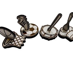 May include: A collection of wooden cutouts featuring sports and culinary themes. Includes a basketball, soccer ball with a shoe, a drum with a drumstick, and a chef's hat with a whisk. Each cutout has a black outline.