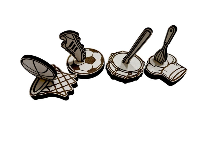May include: A collection of wooden cutouts featuring sports and culinary themes. Includes a basketball, soccer ball with a shoe, a drum with a drumstick, and a chef's hat with a whisk. Each cutout has a black outline.