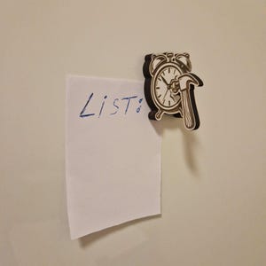 May include: A wooden clock-shaped magnet with a hammer design, holding a white paper with the word "List:" written in blue ink. The magnet is brown and black, and the paper is attached to a white surface.