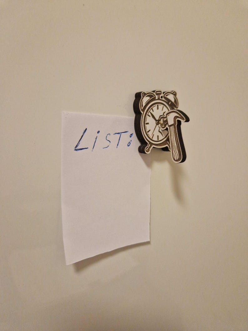 May include: A wooden clock-shaped magnet with a hammer design, holding a white paper with the word "List:" written in blue ink. The magnet is brown and black, and the paper is attached to a white surface.