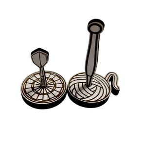 May include: Two wooden decorative items: a dart and a dartboard, and a knitting needle in a ball of yarn. The items are black and white, with a detailed design. The dartboard has a bullseye design.