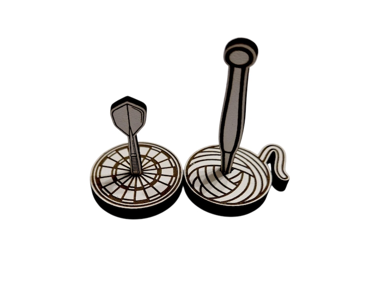 May include: Two wooden decorative items: a dart and a dartboard, and a knitting needle in a ball of yarn. The items are black and white, with a detailed design. The dartboard has a bullseye design.