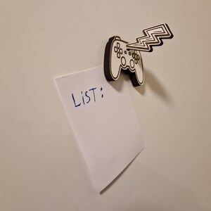 May include: A wooden magnet in the shape of a video game controller with a lightning bolt design. The magnet is holding a white piece of paper with the word "LIST:" written in blue ink. The background is a plain white surface.
