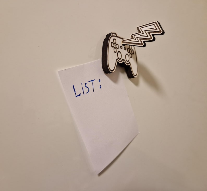 May include: A wooden magnet in the shape of a video game controller with a lightning bolt design. The magnet is holding a white piece of paper with the word "LIST:" written in blue ink. The background is a plain white surface.
