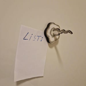 May include: A decorative wooden magnet in the shape of a cloud, holding a small white paper with the word "List:" written in blue ink. The magnet has a black outline and a white interior.