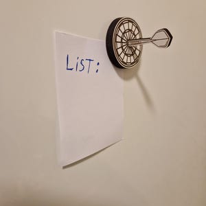 May include: A dartboard-shaped magnet with a dart attached, holding a white piece of paper. The paper has the word "List:" written on it in blue ink. The magnet is black and white.