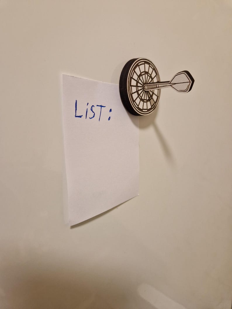 May include: A dartboard-shaped magnet with a dart attached, holding a white piece of paper. The paper has the word "List:" written on it in blue ink. The magnet is black and white.