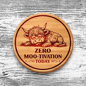 May include: A round bamboo coaster featuring a dark brown engraved illustration of a shaggy Highland cow lying down with its tongue out. Below the cow, the text reads "ZERO MOO-TIVATION TODAY" in a bold, dark brown, sans-serif font.