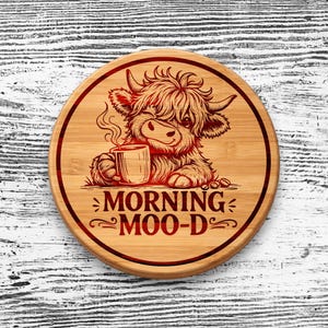 May include: A round bamboo coaster featuring a laser-engraved illustration of a fluffy Highland cow holding a steaming mug. The text MORNING MOO-D is printed below the cow in a bold, dark red font, all enclosed within a circular border.