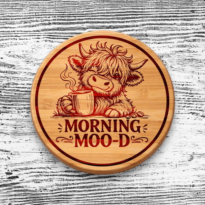 May include: A round bamboo coaster featuring a laser-engraved illustration of a fluffy Highland cow holding a steaming mug. The text MORNING MOO-D is printed below the cow in a bold, dark red font, all enclosed within a circular border.