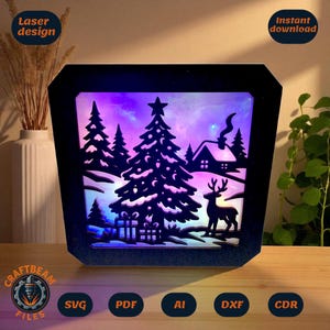 Puede incluir: Una escena navideña negra cortada con láser con un árbol, un ciervo y una casa, iluminada por una luz púrpura y azul. El marco tiene una forma geométrica. Se ven las palabras "Laser design" e "Instant download".