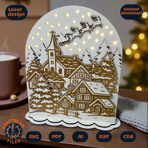 Op de afbeelding: Een witte lasergesneden kerstdorp-scène met een kerk, huizen en bomen. Het ontwerp bevat de slee en rendieren van de kerstman. De woorden "Laser design" en "Instant download" zijn zichtbaar.