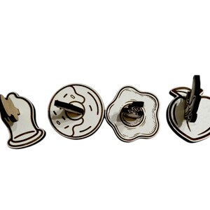 May include: Four wooden food-themed decorative items: a bell, a donut, a fried egg, and a peach. Each item is made of light-colored wood with dark brown outlines and accents. The donut has a hole and the egg has a yolk.
