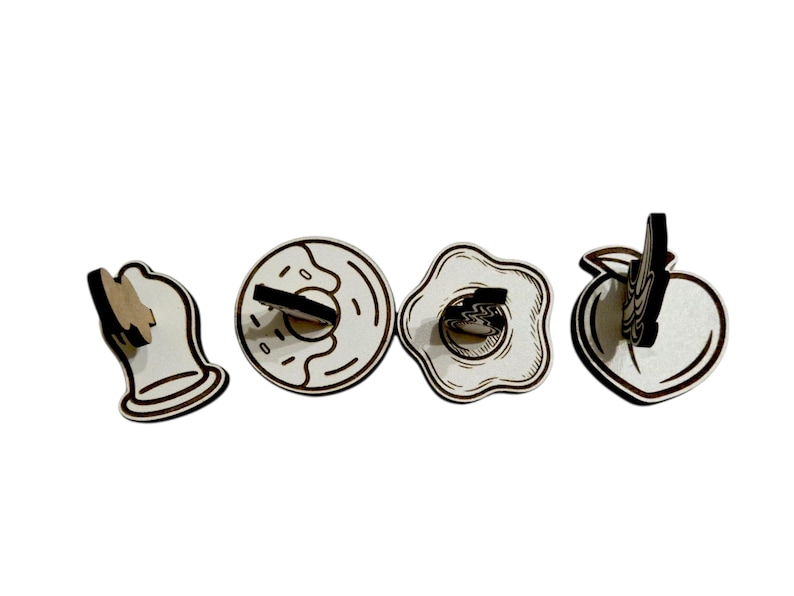 May include: Four wooden food-themed decorative items: a bell, a donut, a fried egg, and a peach. Each item is made of light-colored wood with dark brown outlines and accents. The donut has a hole and the egg has a yolk.