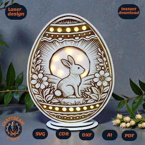 Op de afbeelding: Een lasergesneden houten paasei-decoratie met een konijn, bloemen en een zonnestraalontwerp. Het eivormige licht heeft een witte basis en wordt verlicht met kleine lampjes. De tekst "Laser design" en "Instant download" zijn zichtbaar.