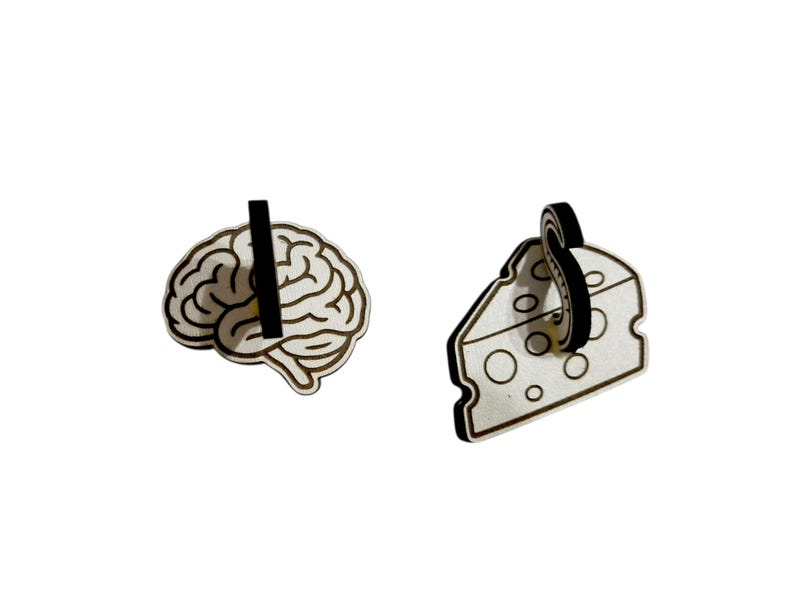 May include: Two wooden earrings: one shaped like a brain and the other like a slice of cheese. Both earrings have a black post and are light brown with black outlines. The brain earring has a vertical black line through it.