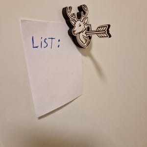 May include: A wooden magnet featuring a deer head with antlers, an arrow, and a shield design. The magnet holds a white piece of paper with the word "LIST:" written in blue ink. The magnet is brown and white.