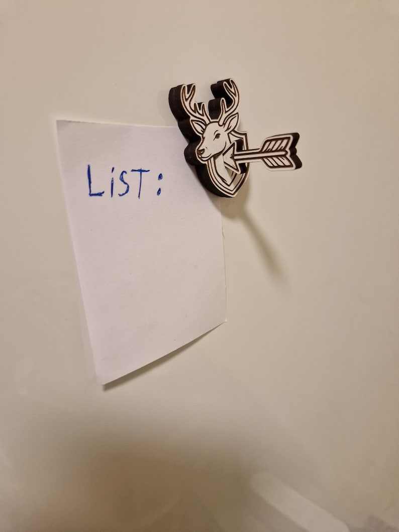 May include: A wooden magnet featuring a deer head with antlers, an arrow, and a shield design. The magnet holds a white piece of paper with the word "LIST:" written in blue ink. The magnet is brown and white.