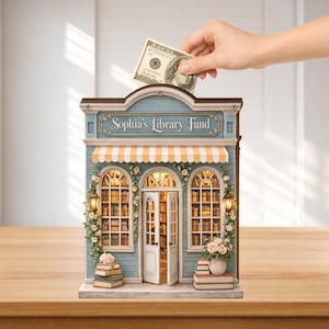 May include: A blue and brown wooden bank shaped like a library, with the text "Sophia's Library Fund". The bank has a yellow and white striped awning, detailed windows, and a stack of books. A hand is inserting a $100 bill into the slot.