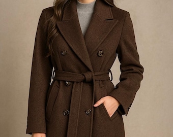 Könnte beinhalten: Ein langer, doppelreihiger brauner Mantel mit passendem Gürtel. Der Mantel hat ein fallendes Revers, zwei Knopfreihen und Taschen. Das Model trägt einen grauen Rollkragenpullover darunter.