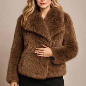 May include: A brown faux fur coat with a wide collar and long sleeves. The coat is knee-length and has a soft, textured appearance. The model is wearing a black skirt.