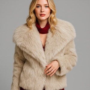 May include: A beige faux fur jacket with a wide collar. The jacket is open, revealing a red top underneath. The model has blonde hair and is wearing a red scarf. The jacket is a neutral color.