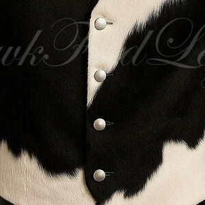 May include: A vest featuring a black and white cowhide pattern with a textured, fur-like appearance. The garment includes a front button closure with four round, silver-toned metallic buttons aligned vertically along the center placket.