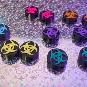 May include: A collection of black cylindrical objects, each topped with a colorful biohazard symbol in shades of blue, pink, orange, yellow, and teal. The objects are arranged on a shimmering, star-patterned surface.