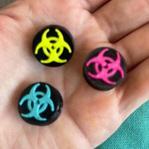 May include: Three black round buttons with a biohazard symbol in neon yellow, hot pink, and teal. The buttons are small and appear to be made of a glossy material. The biohazard symbols are centered on each button.